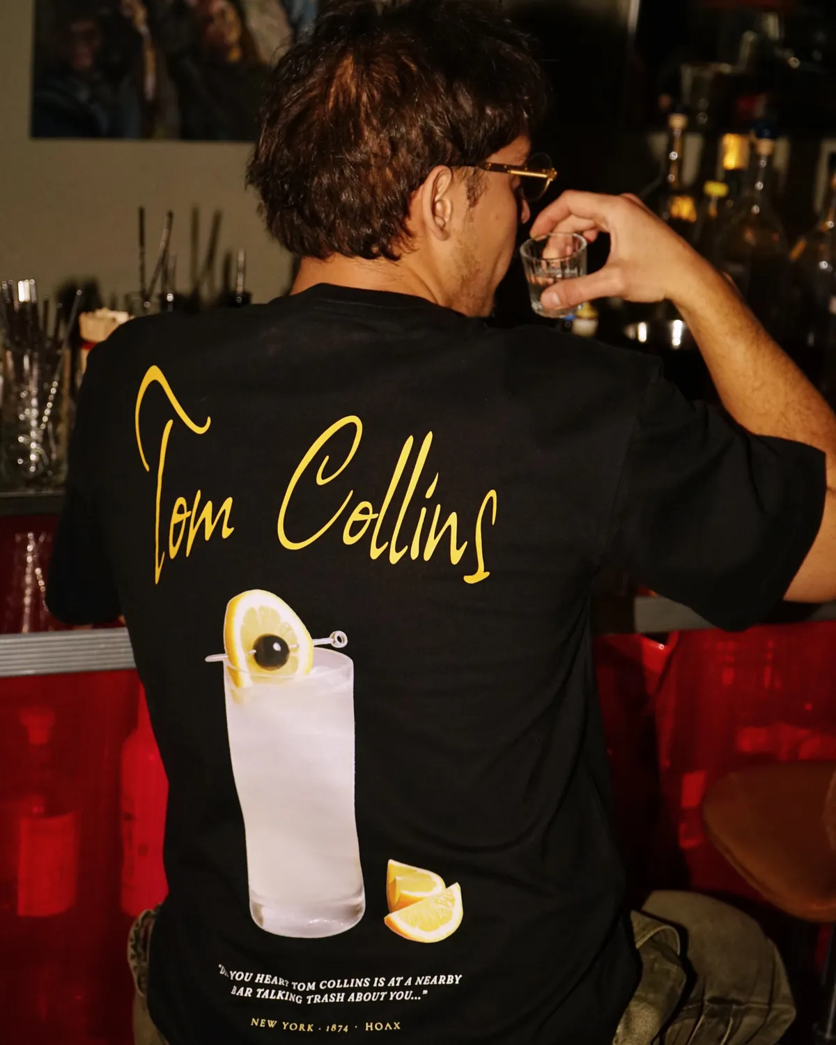Persona de espaldas en un bar con la playera Tom Collins, sosteniendo un vaso
